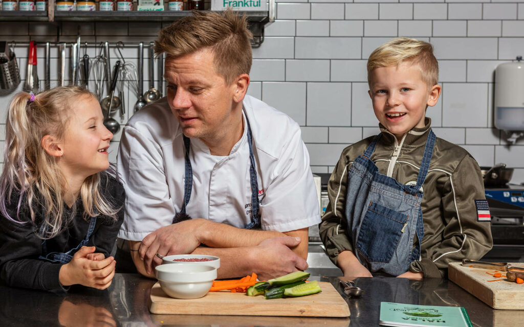 Kocken Marcus hänger mot bänken i ett kök och pratar med två barn som står på varsin sida om honom.